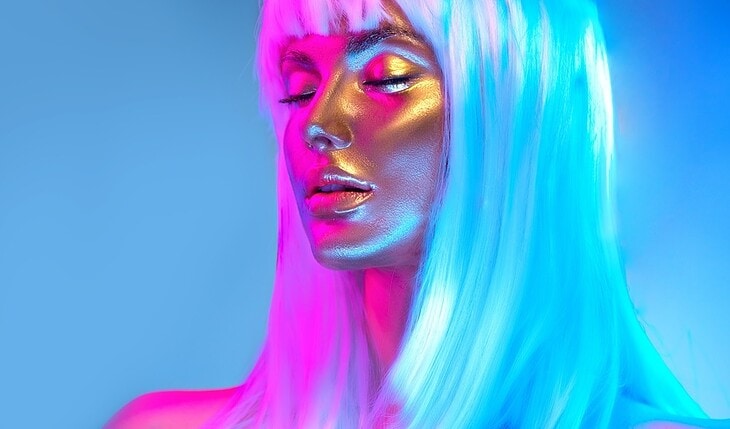 Glowing gold skin. Fashion model woman face in bright neon colourful lights, beautiful sexy woman with white hair and trendy make-up. Art design make up. Glittering, golden, metallic shine makeup